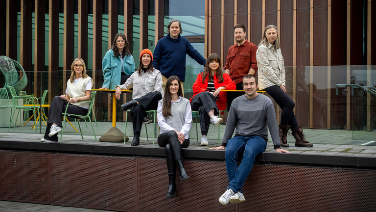 A group of ten people pose together outdoors on a modern terrace with metal and glass architectural elements in the background. They are dressed in casual, contemporary clothing.